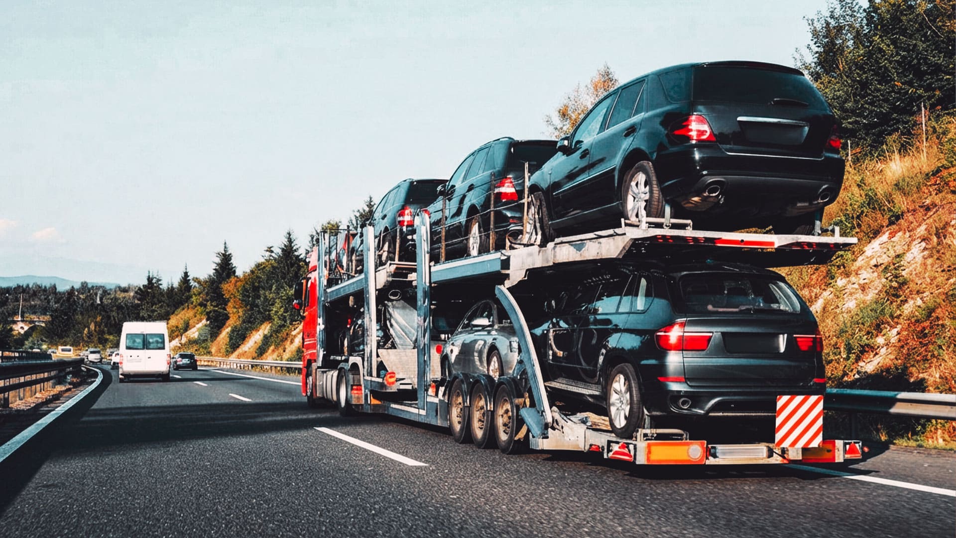 Camión nodriza o portacoches de gran tamaño circulando por una carretera abierta, transportando varios vehículos oscuros de importación.